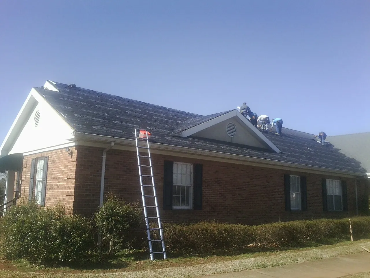 Crew tearing shingles off a brick ranch roof with a ladder set — Metal Roof Repair work by our crew in Wittman