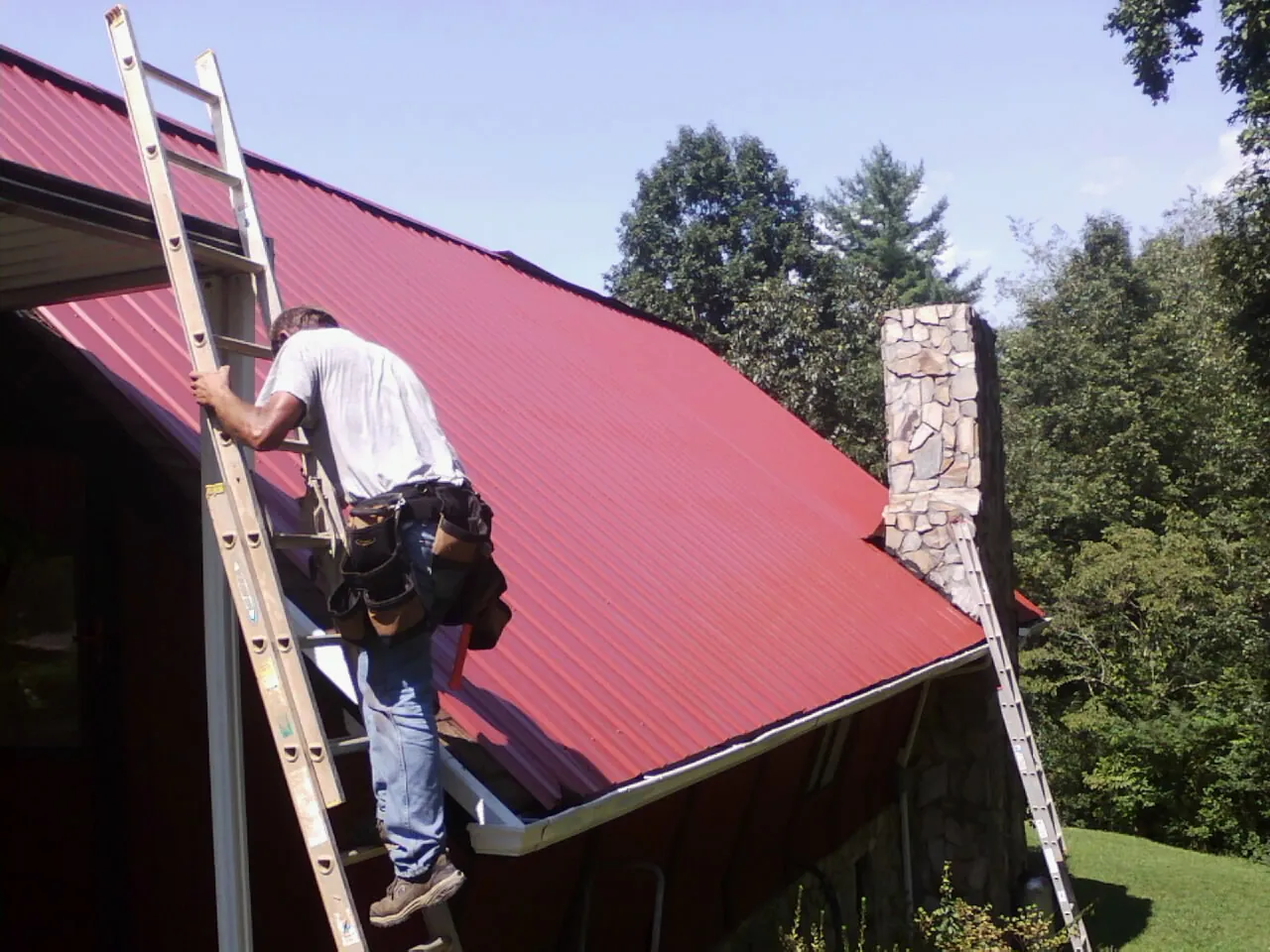 Roofer on a ladder beside a red metal roof and stone chimney — Soffit & Fascia Repair by Roofing Brothers in Wittman